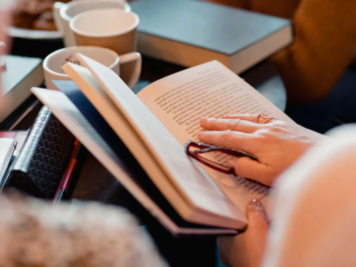 Une personne tient un livre ouvert sur une table couverte de livres et de tasses.