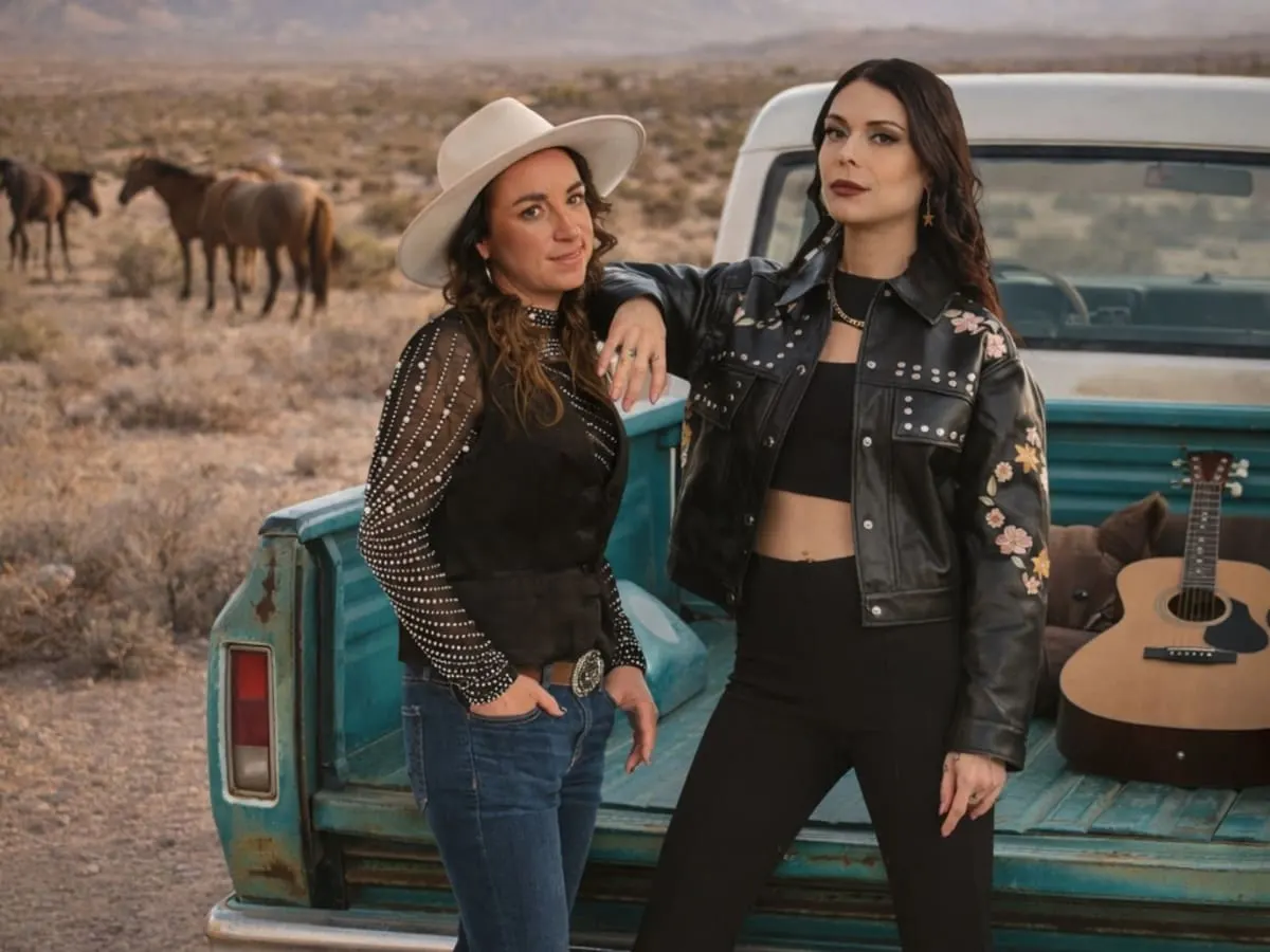 Deux femmes habillées en noir posent devant une camionnette où l’on aperçoit une guitare. En arrière-plan, un paysage typique du Far West, avec des chevaux.