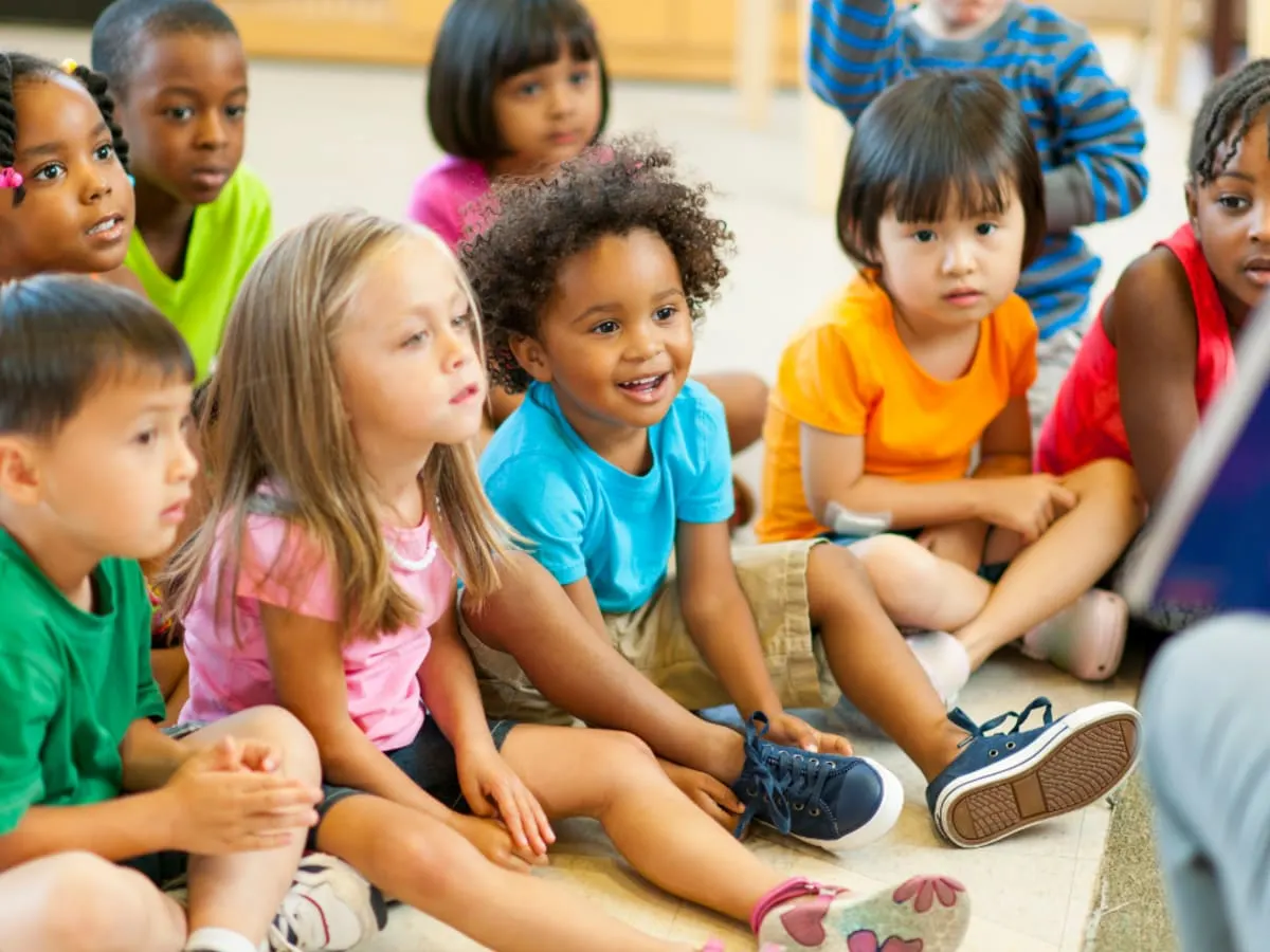 Un groupe d’enfants assis sur un tapis pendant une activité de lecture, qui regardent un livre présenté par une personne hors champ.