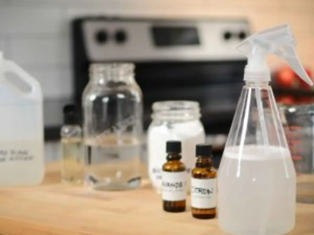 Sur une table de cuisine sont posés : un vaporisateur transparent avec un liquide blanchâtre, deux flacons d'huiles essentielles, 3 pots de verre remplis et une cruche, peut-être de vinaigre. Il y a une cuisinière à l'arrière-plan.