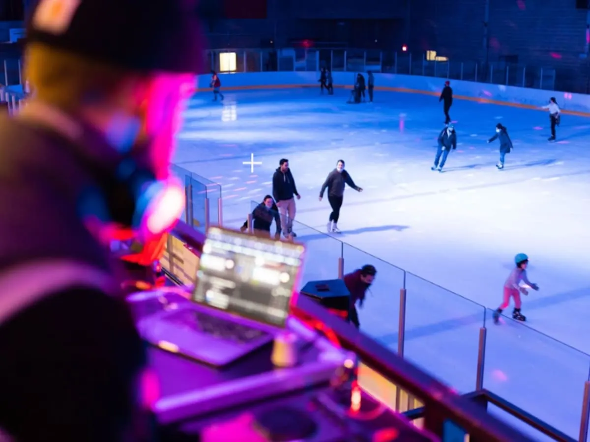 Une patinoire éclairée de lumières colorées où plusieurs personnes patinent avec à l'avant-plan une personne à une console de DJ.
