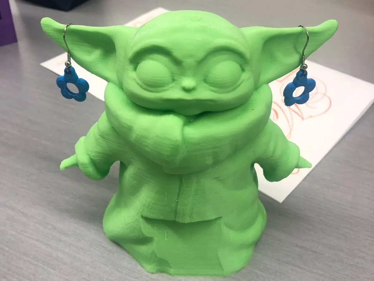 Photo d'un petit Yoda imprimé en 3D portant des boucles d'oreille en fleur créées également grâce à l'impression 3D.