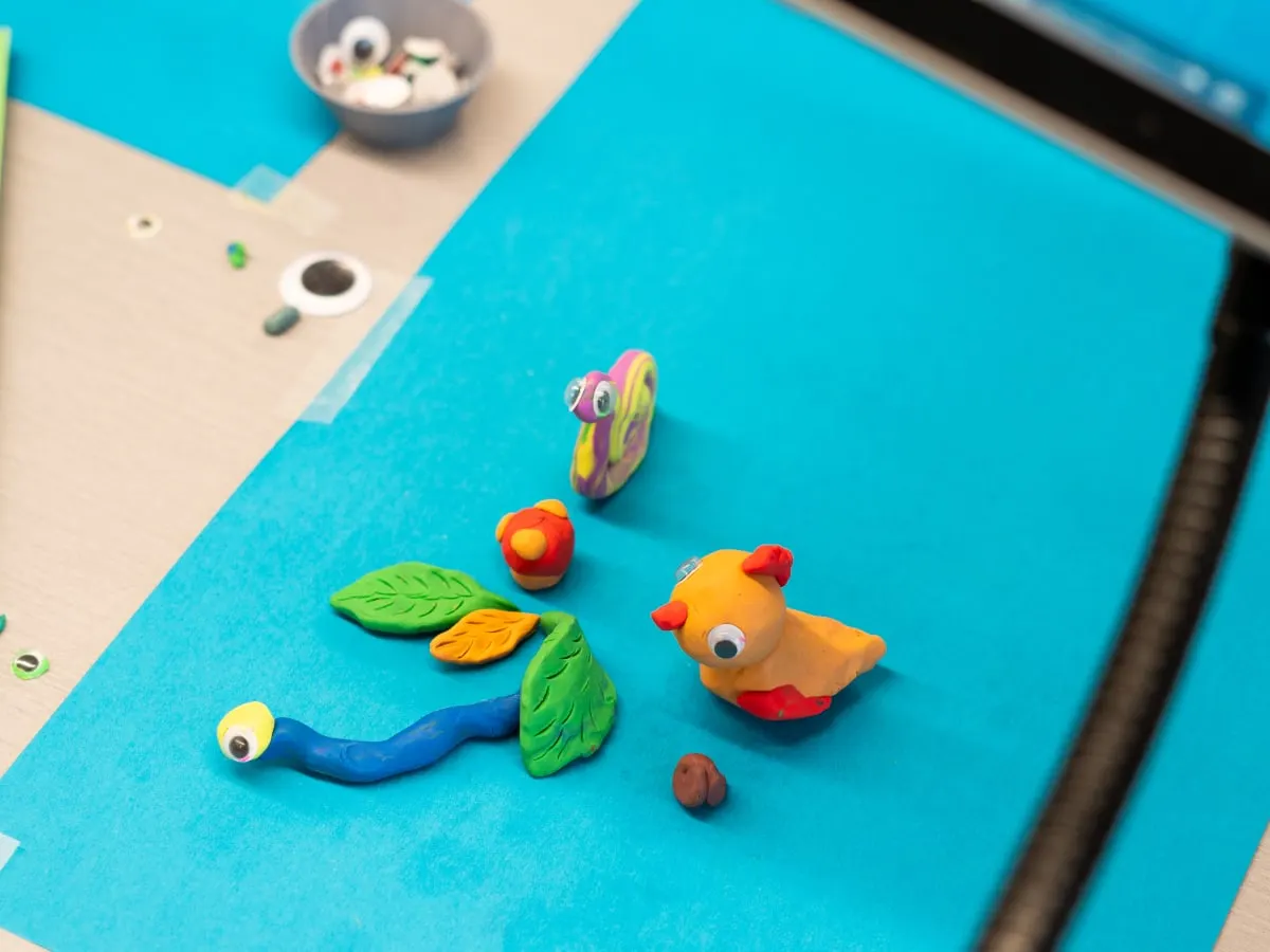 des personnages en pâte à modelée sont disposés sur un fond bleu et une tablette dans le coin de l'image indique l'application d'animation image par image utilisée dans l'activité